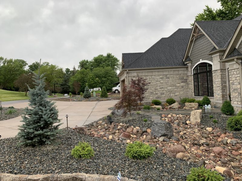 Stone house with gray roof, blue spruce, and rock landscaping under an overcast sky.