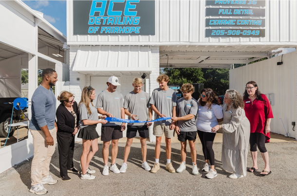 People cutting a ribbon at the grand opening of Ace Detailing of Monroe, outdoor shop front, sunny day.