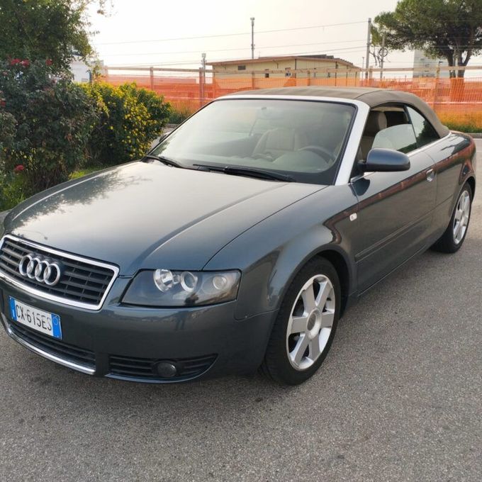Audi A4 decappottabile grigia parcheggiata sul marciapiede; cerchi argentati, interni color cuoio, capote alzata.