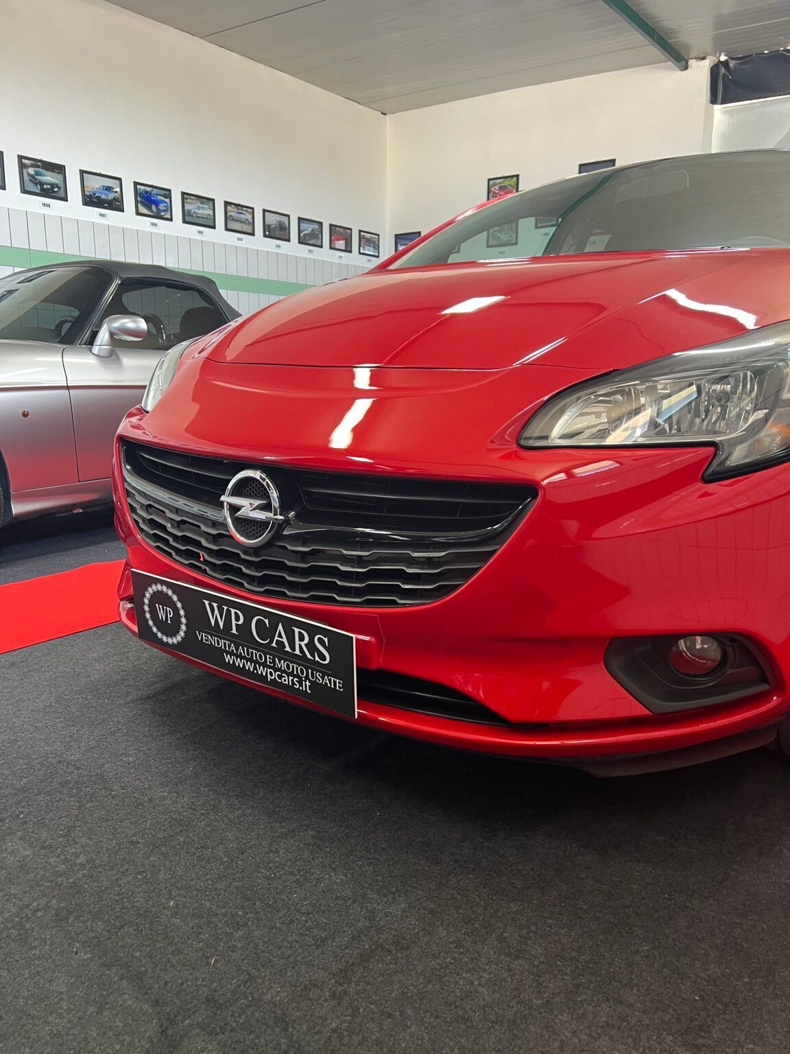 Opel Corsa rossa parcheggiata all'interno di uno showroom; griglia nera, insegna WP CARS, auto argentata accanto.