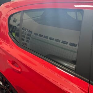 Lato dell'auto rossa con finestrino che riflette una stanza con più display incorniciati.