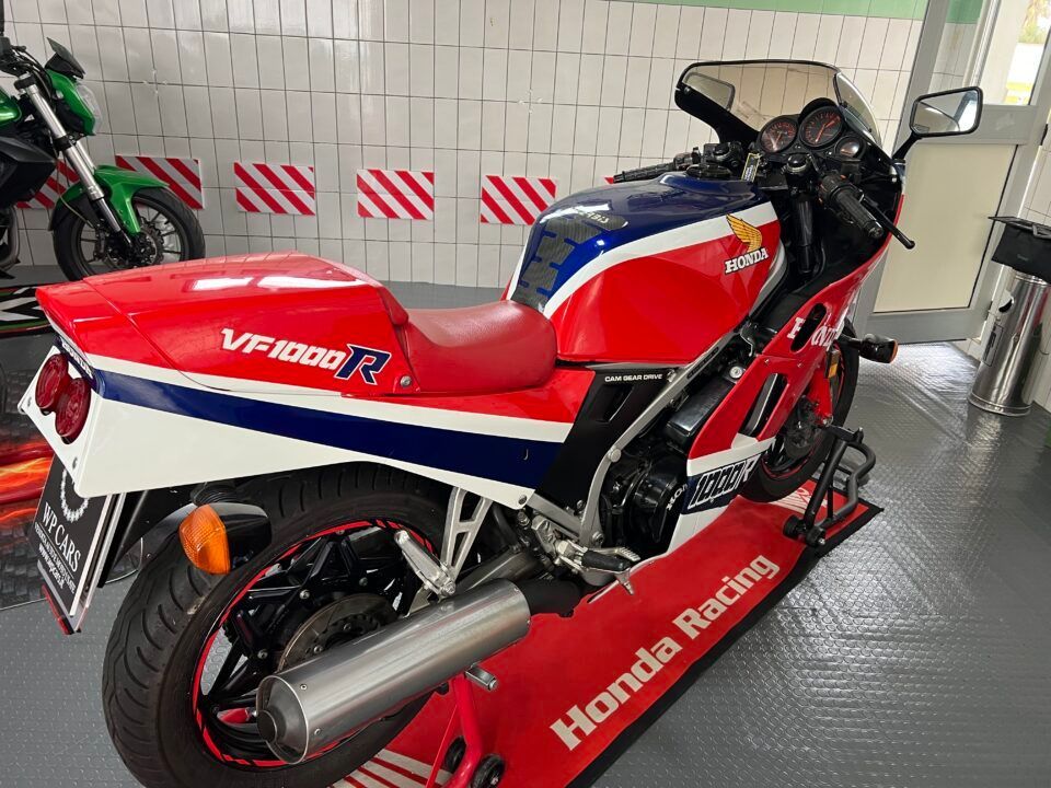 Motocicletta Honda VF1000R rossa, bianca e blu su un supporto in un garage, con una motocicletta verde sullo sfondo.