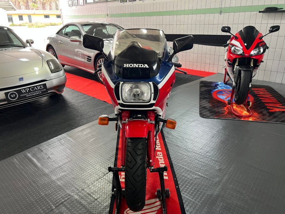 Motocicletta Honda rossa, bianca e blu esposta in un garage, con altri veicoli visibili.