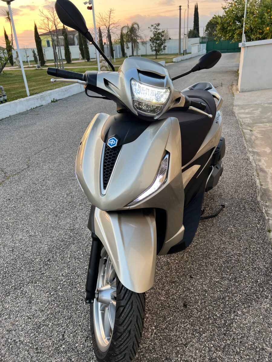 Uno scooter Piaggio argentato parcheggiato su un vialetto asfaltato, con il sole al tramonto sullo sfondo.