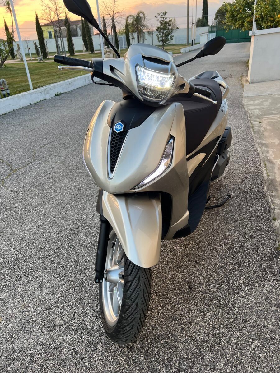 Uno scooter Piaggio argentato parcheggiato su un vialetto asfaltato, rivolto verso l'osservatore. Di giorno.