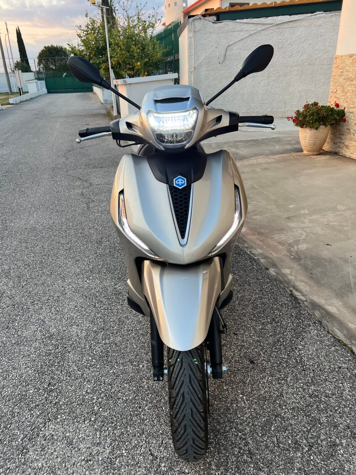 Uno scooter argentato parcheggiato su una strada asfaltata. Ha un faro LED bianco e un sedile nero, con sfondo sfocato.