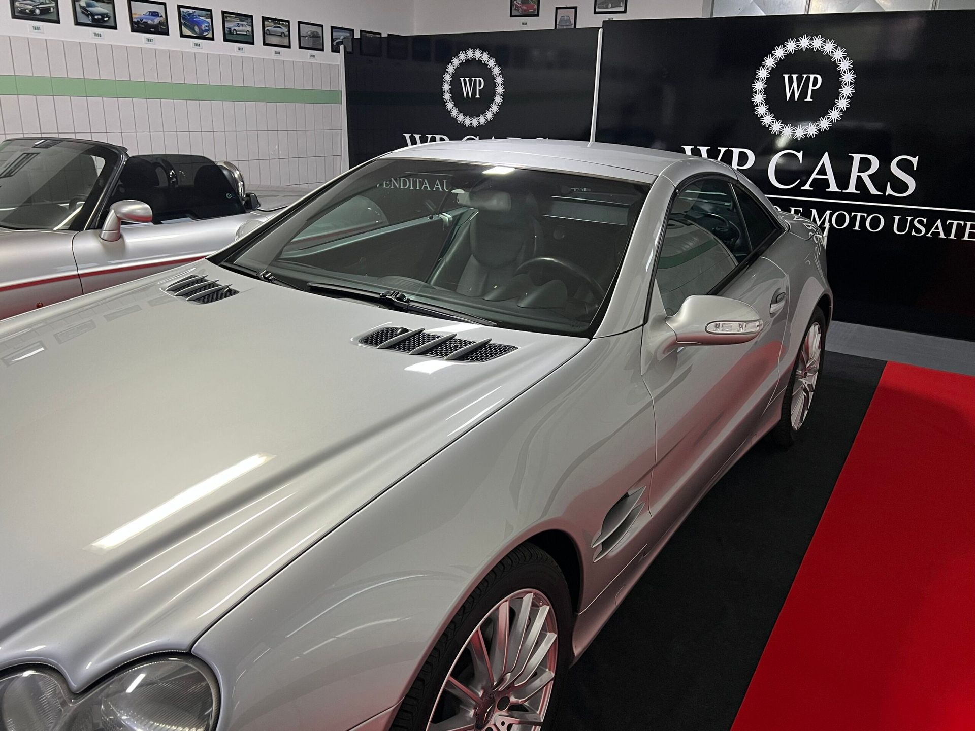Una Mercedes-Benz Classe SL coupé argentata in uno showroom, parcheggiata su un tappetino rosso e nero.