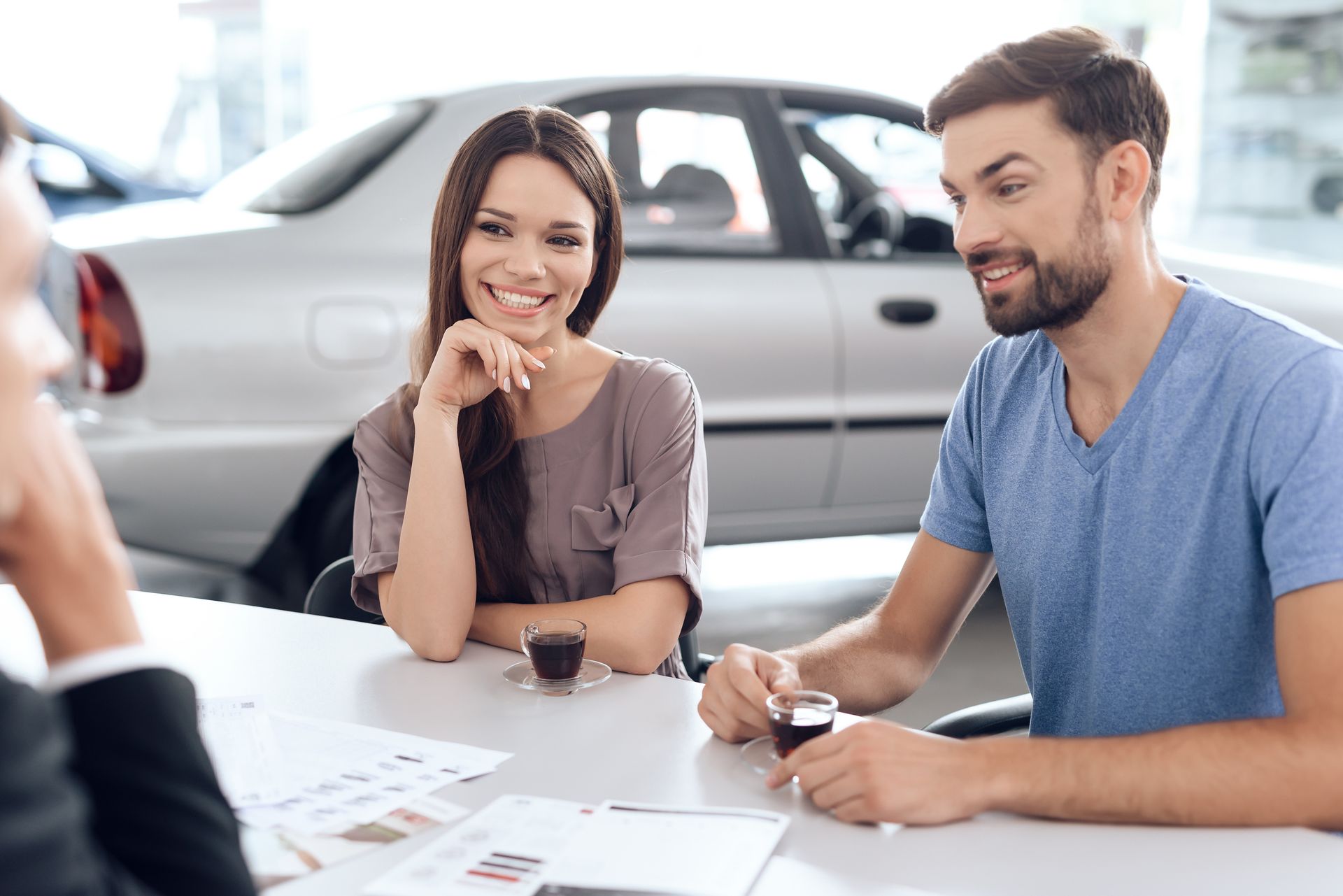 Coppia sorridente al tavolo di una concessionaria di automobili, con il venditore e un'auto sullo sfondo.