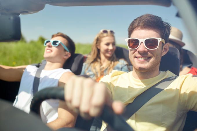 People in a convertible car, driving on a sunny day. Driver wearing sunglasses smiles. Passengers appear happy.