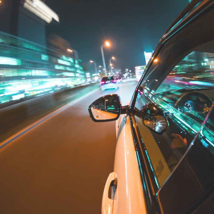Car speeding at night, blurred lights, reflection in side mirror, city background.