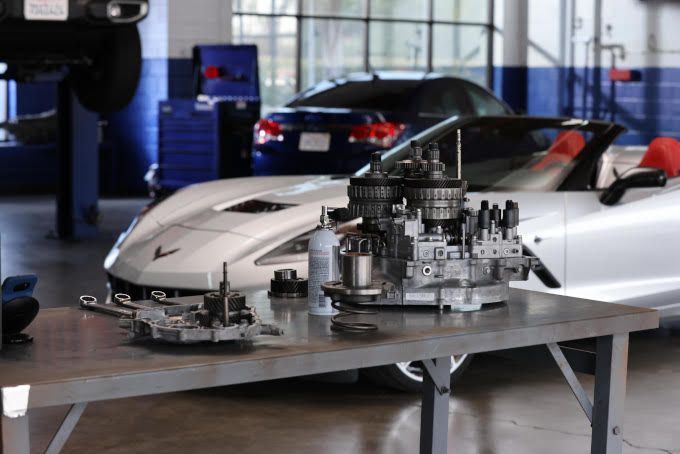 Disassembled car transmission parts on a table in a garage, with a silver Corvette in the background.