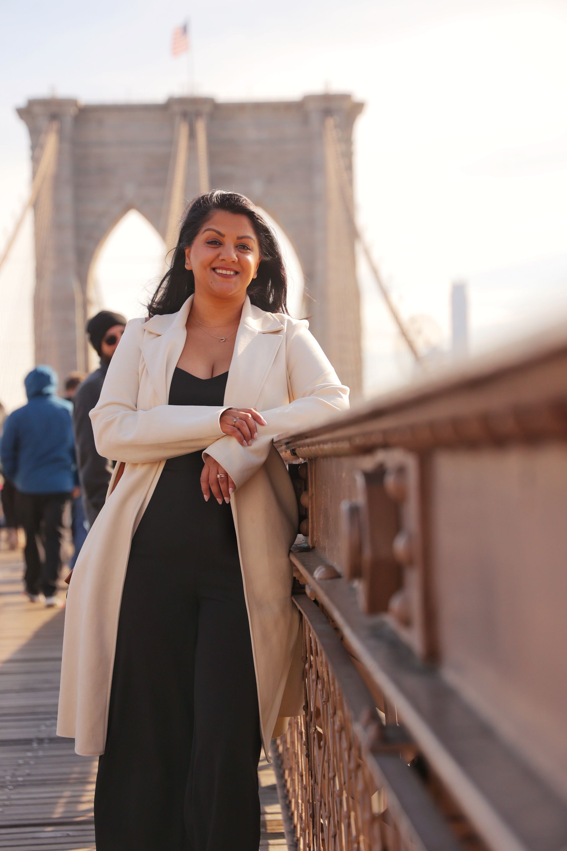 A smiling person in a cream coat and black jumpsuit stands leaning against the railing of the Brooklyn Bridge.