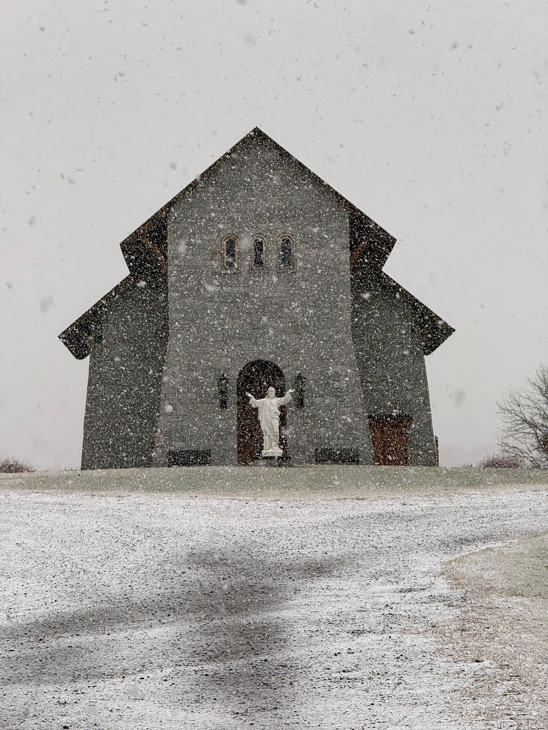 A stone church with a statue of a figure with outstretched arms in the entrance, standing in a snowy field during a snowfall.