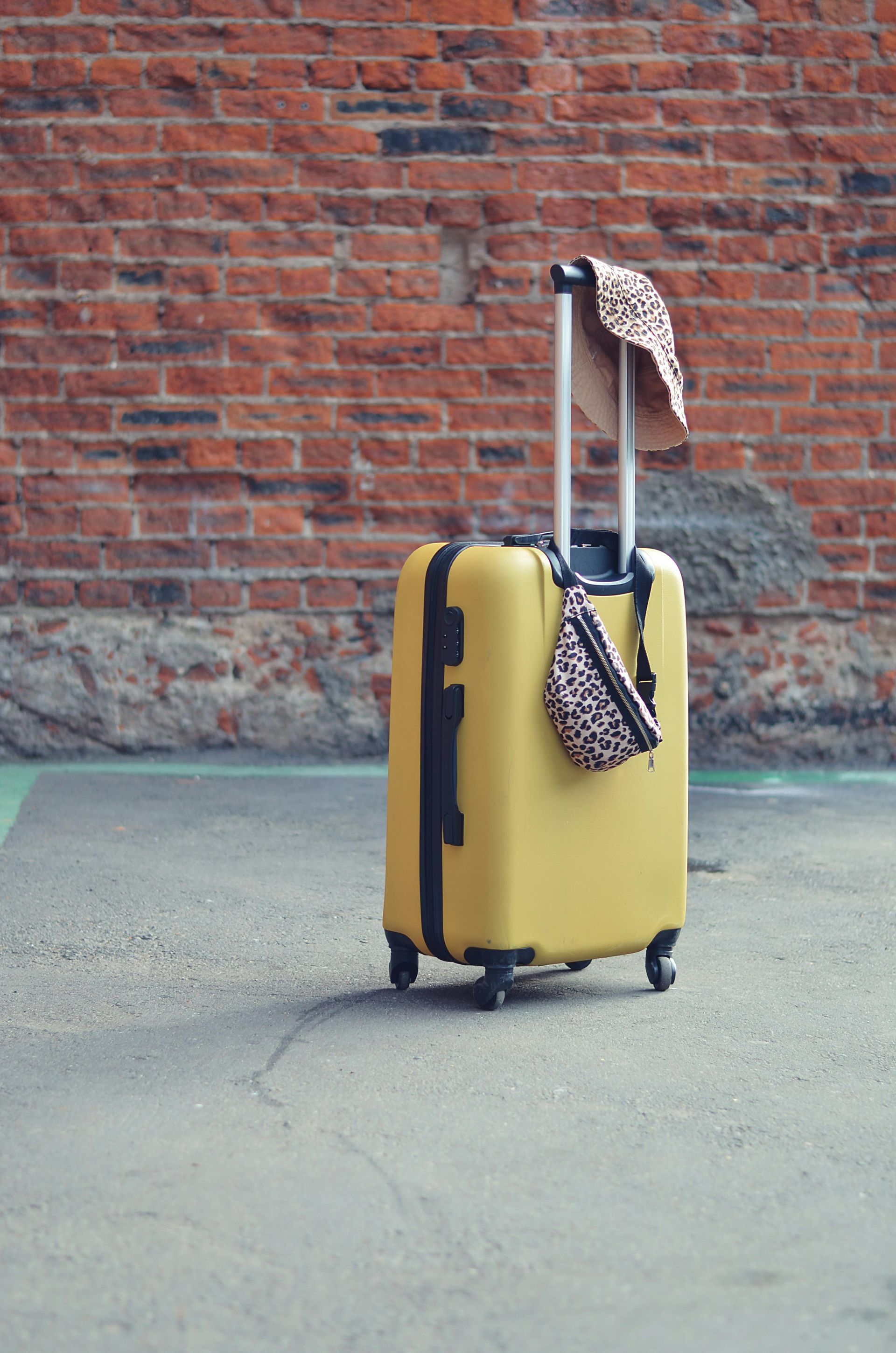 A yellow suitcase stands on a gray concrete surface in front of a red brick wall, with a leopard-print bag hanging on it.