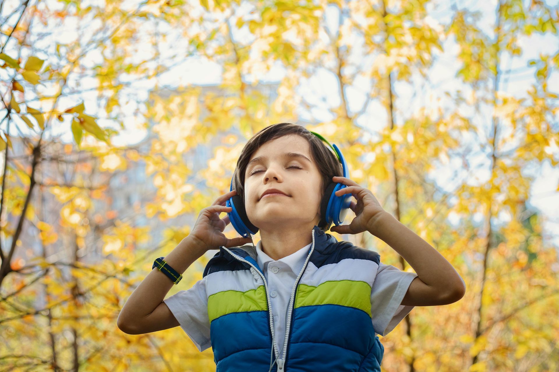 A person with eyes closed wearing blue headphones and a colorful vest outdoors, surrounded by yellow autumn trees.