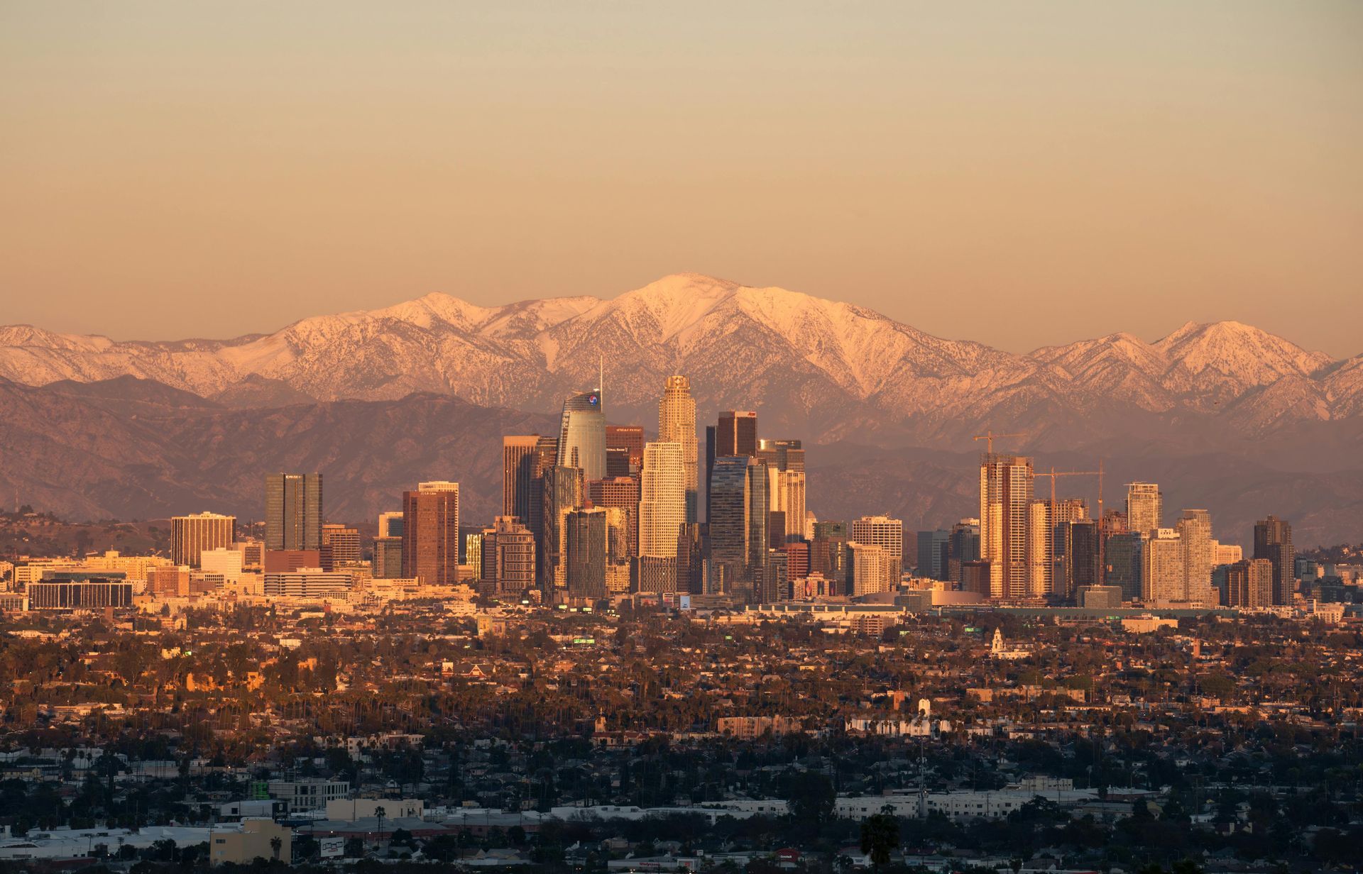 A golden-hour shot of the Los Angeles skyline with snow-capped mountains in the background.
