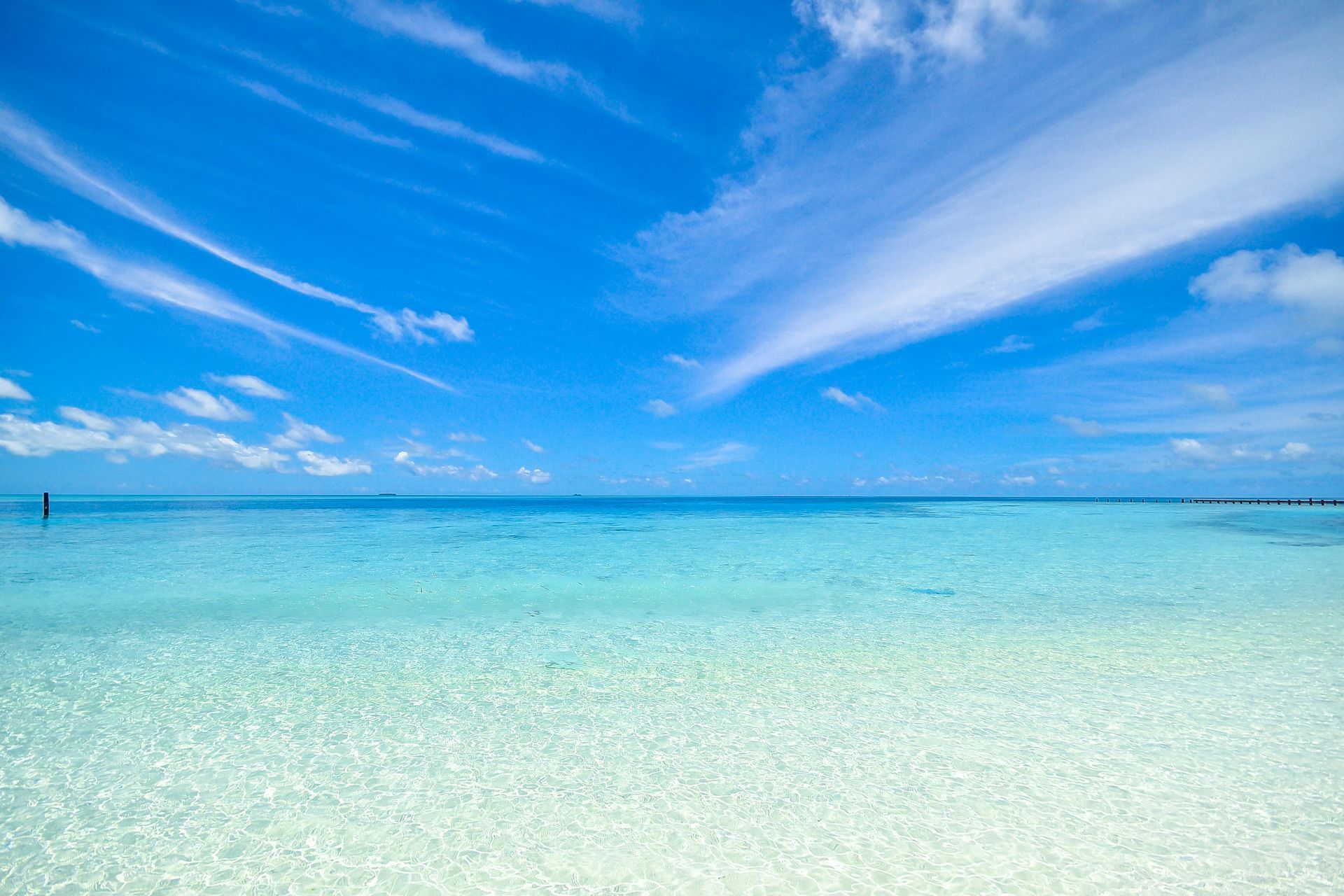 A tranquil tropical beach with crystal-clear turquoise water stretching to the horizon under a bright blue, cloudy sky.