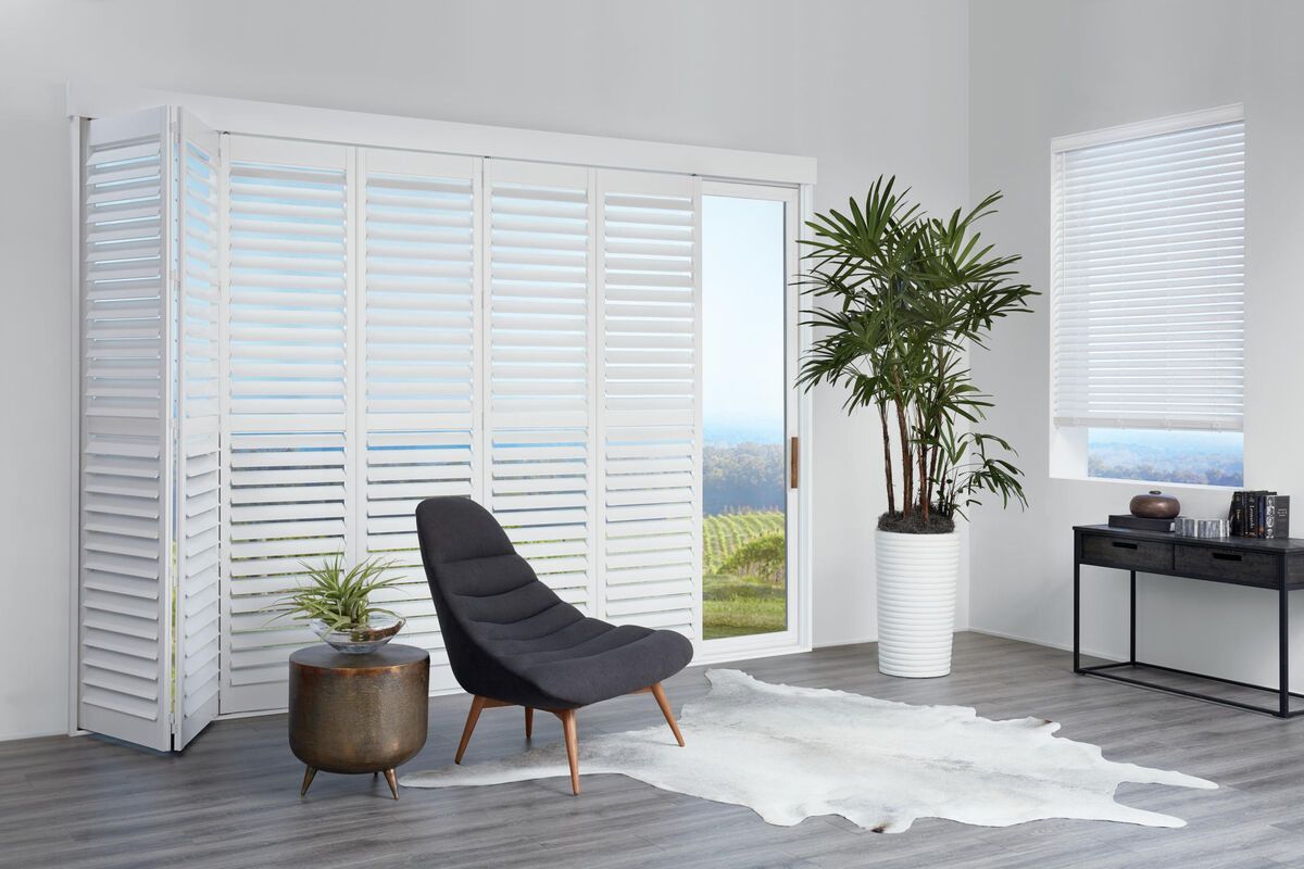 A living room with white shutters , a chair , and a plant.