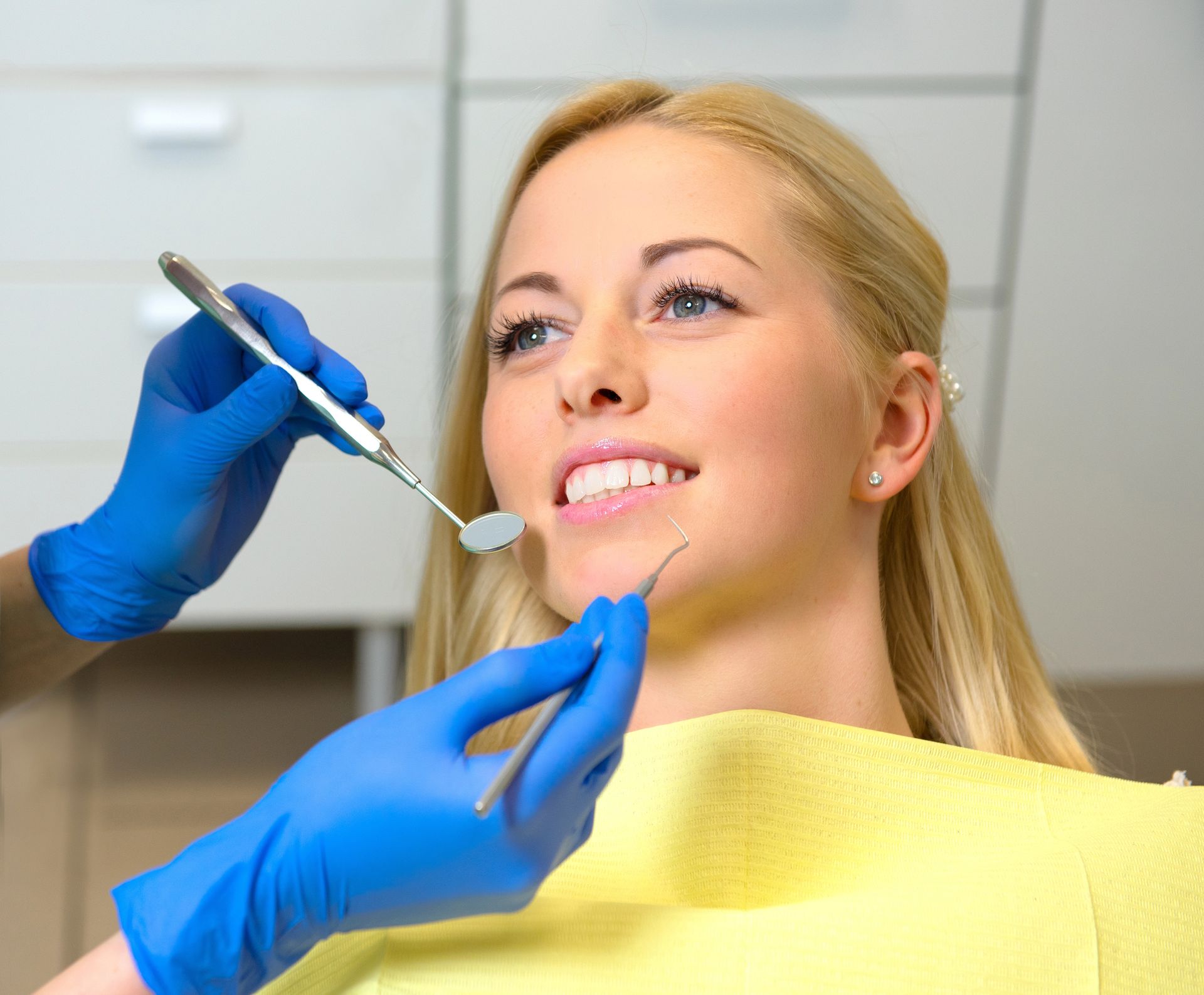 Un dentista con i guanti blu utilizza uno specchietto e uno strumento odontoiatrico per esaminare un paziente sorridente seduto sulla poltrona del dentista.