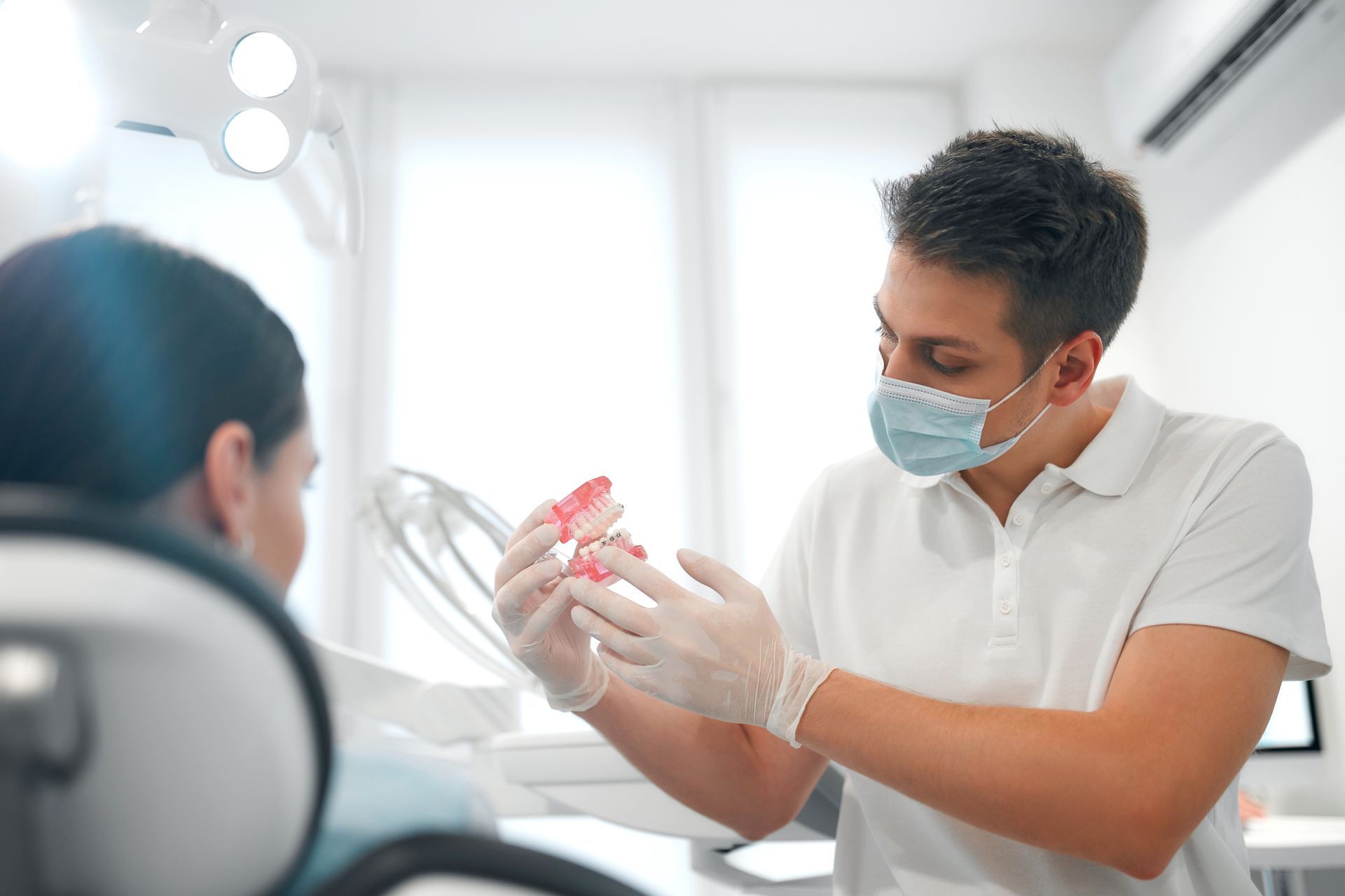 Un dentista, con indosso mascherina e guanti, tiene in mano un modellino dentale per spiegare una procedura a un paziente in uno studio medico.
