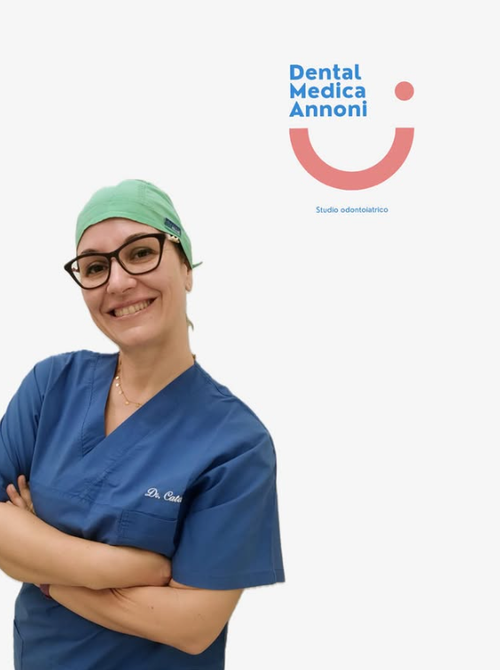 Un operatore sanitario sorridente, in camice blu e cuffia verde, posa davanti a uno sfondo bianco con il logo di uno studio dentistico.