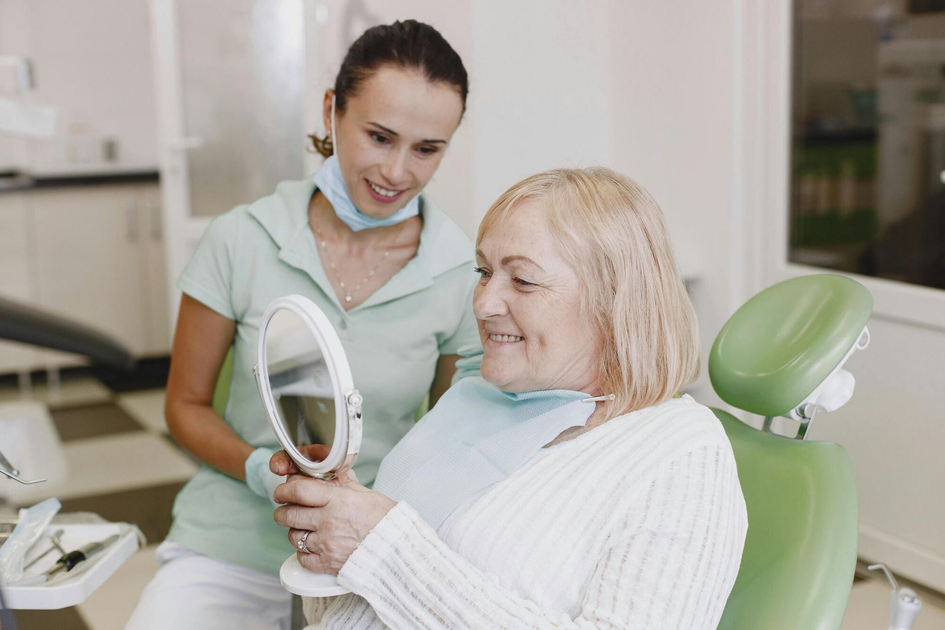 A woman is sitting in a dental chair looking at her teeth in a mirror.