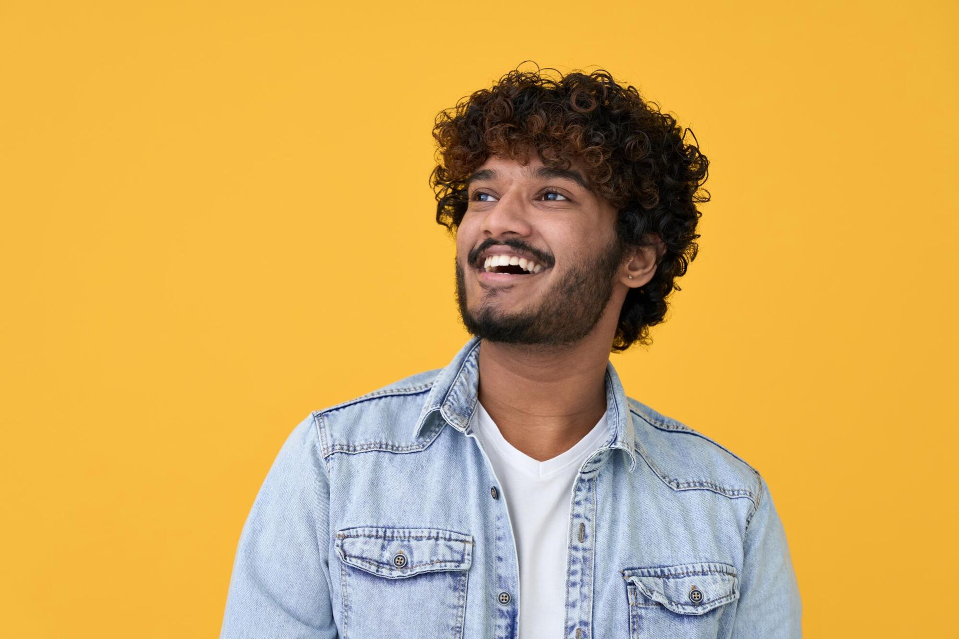 Man with curly brown hair and a beard, smiling while looking upwards against a yellow backdrop, wearing a denim jacket.