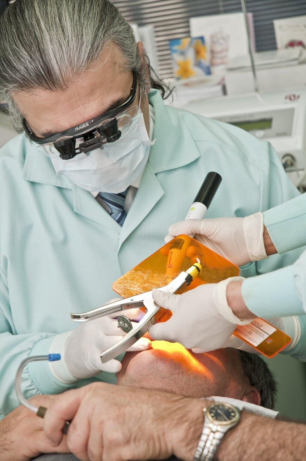 Dentist performing dental work on a patient in a clinic. Tools visible. Patient is lying down.