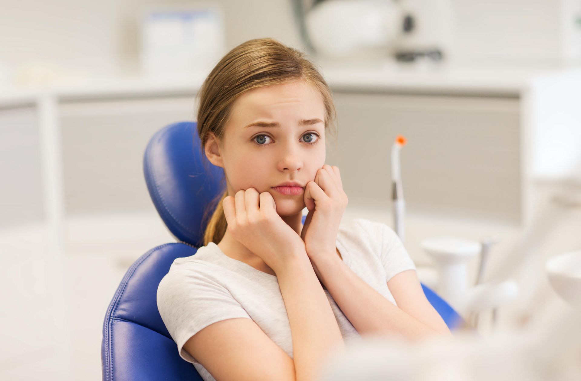 Person in a dental chair, looking worried, hands on cheeks. Brightly lit dental office in background.