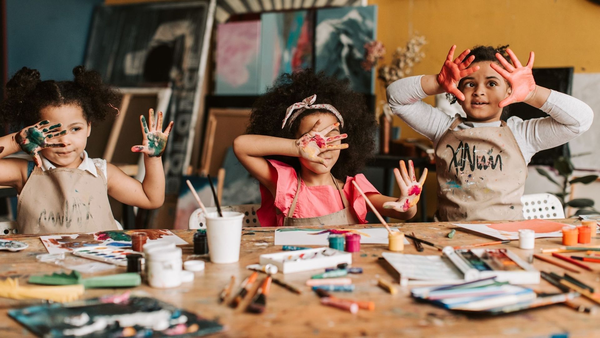 Three children with paint-covered hands and faces at a messy art table, posing with creative expressions.