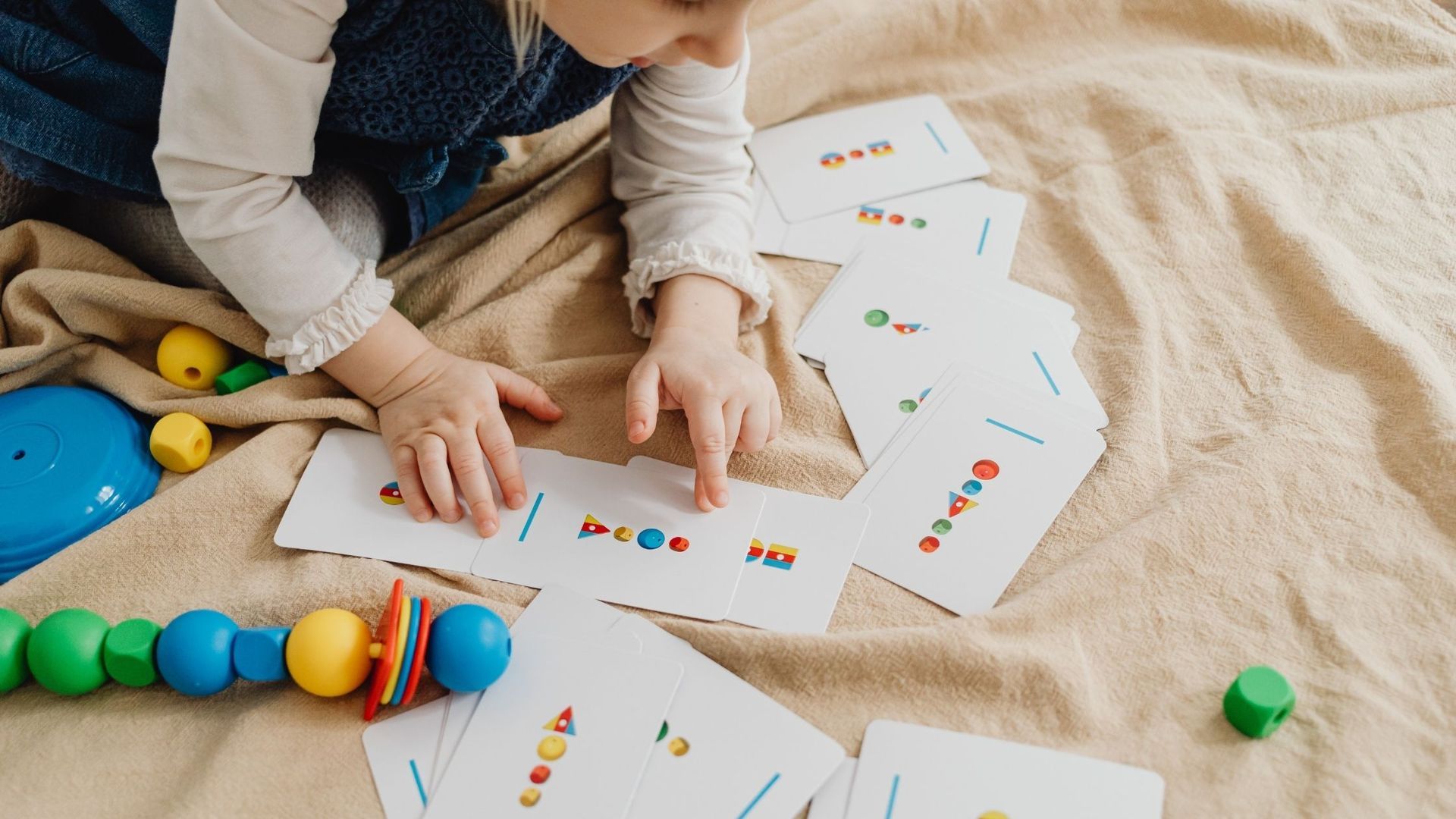 Child pointing to a card with colorful circles on it, surrounded by similar cards and toys on a blanket.
