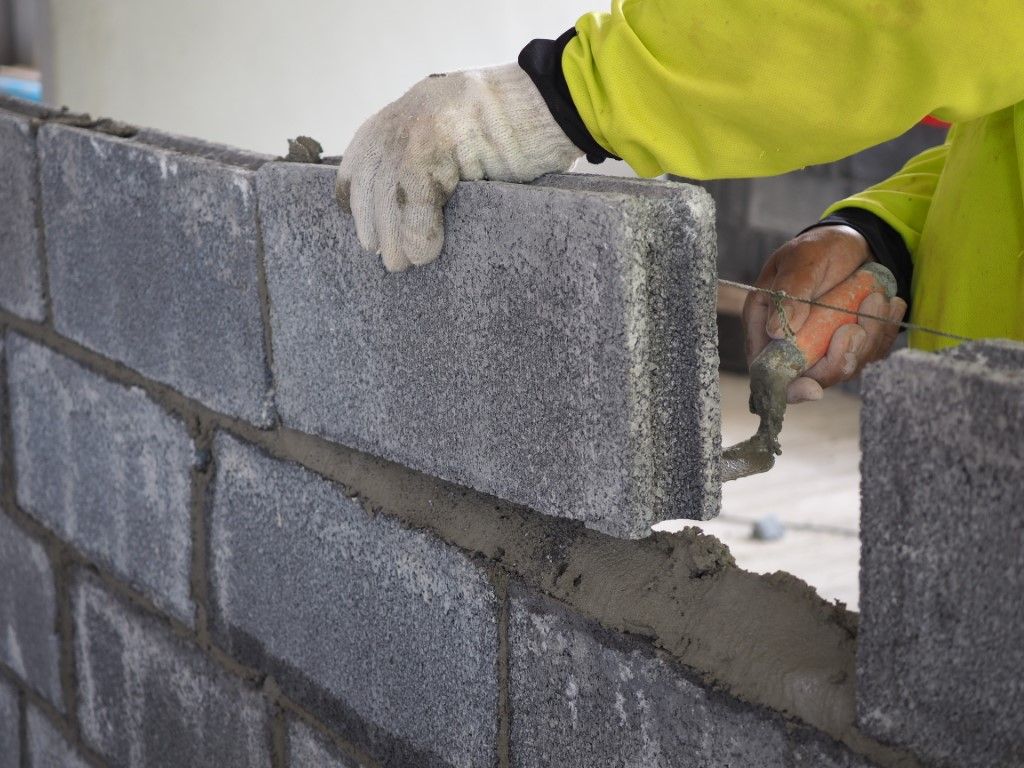 Construction worker laying concrete blocks on a wall, wearing a yellow jacket and gloves.
