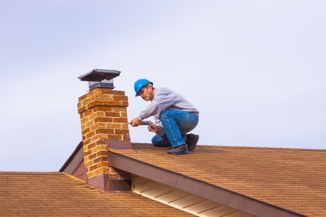 Worker in a blue hard hat repairing a brick chimney on a shingled roof