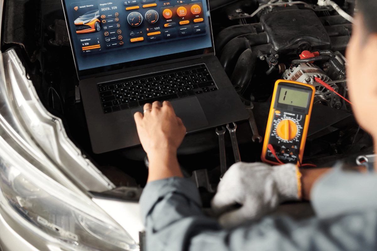A man is working on a car with a laptop and a multimeter.