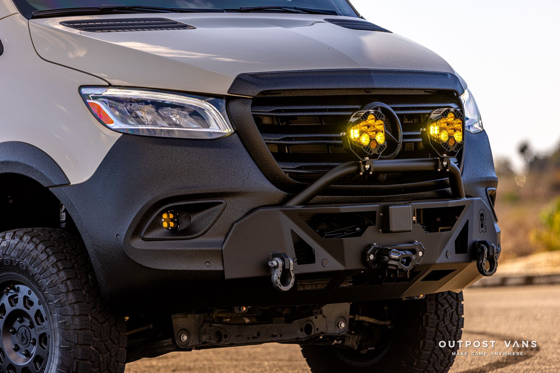 A white van with a black bumper and yellow lights is parked on the side of the road.