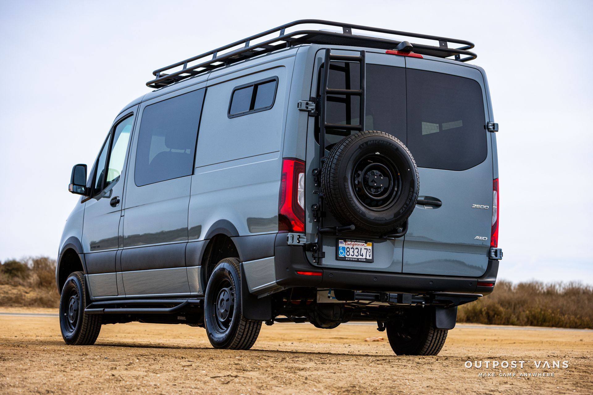 A gray van with a ladder on the back is parked in a dirt field.