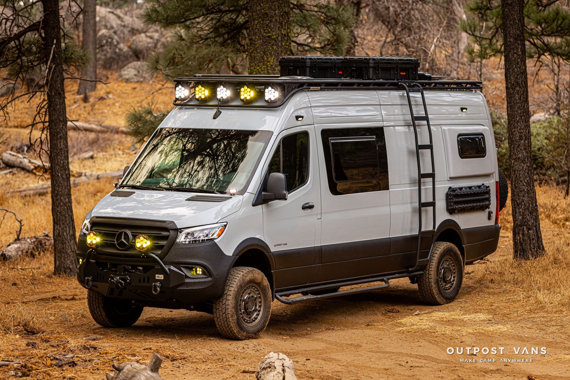 A white van with a ladder on the roof is parked in the woods.