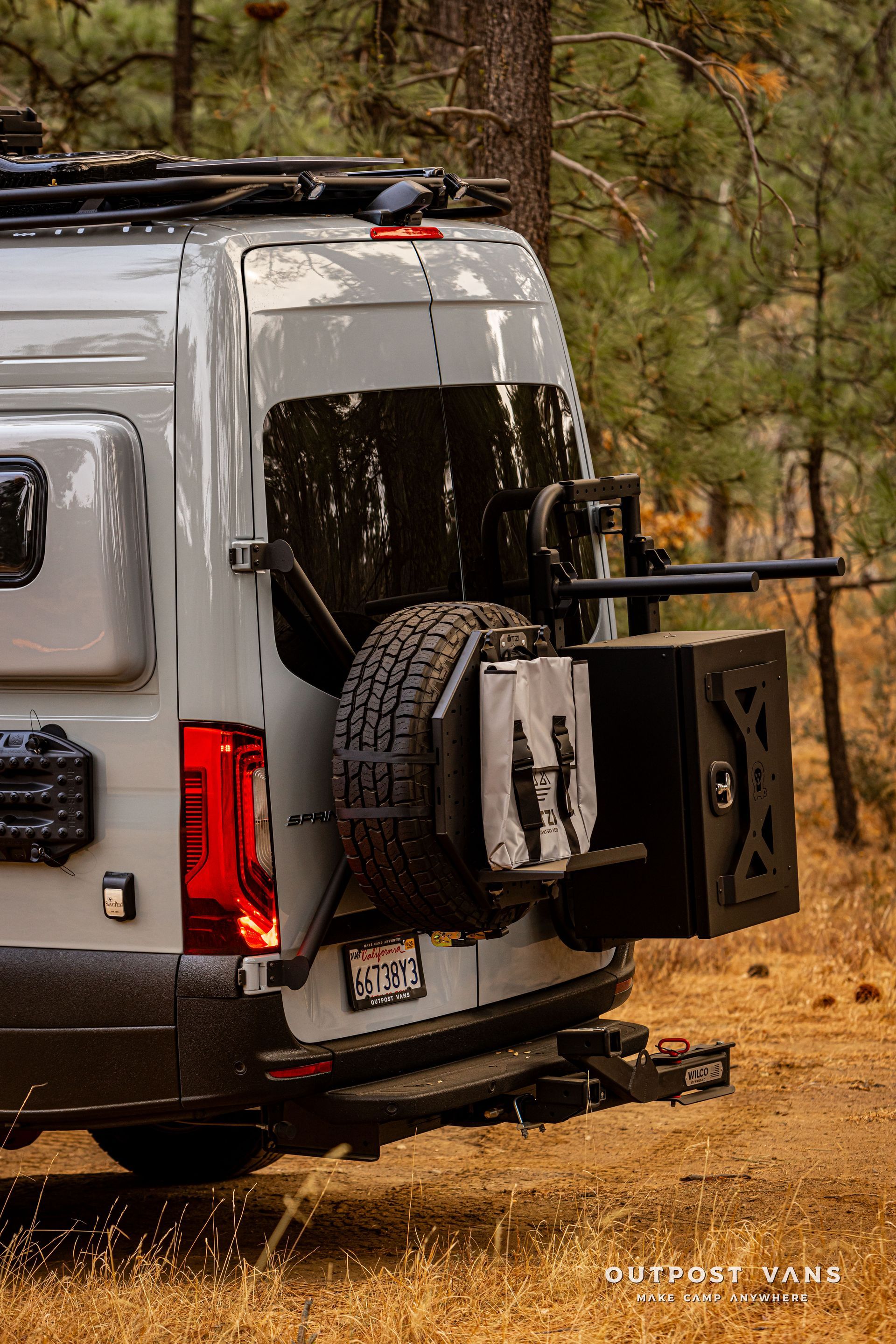 A van with a spare tire attached to the back is parked in the woods.