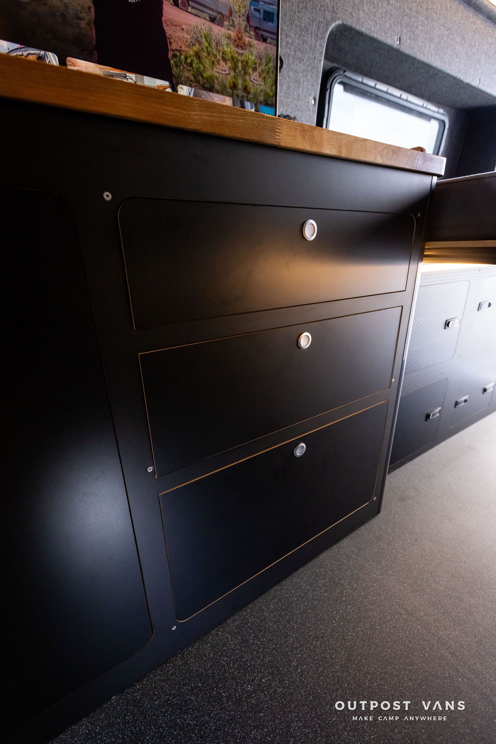 Outpost Vans custom built camper van Ford Transit 148 AWD. A black cabinet with drawers and a wooden counter top in a van.