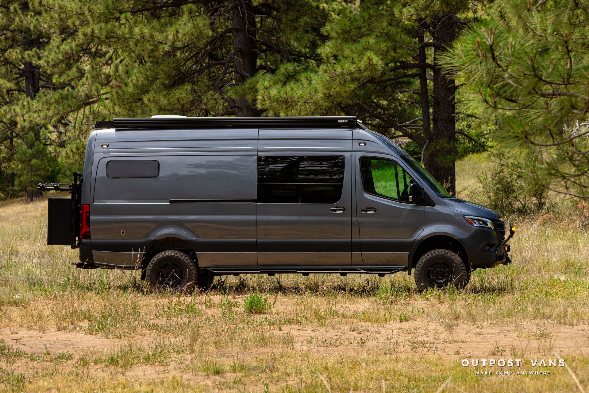 A gray van is parked in a field with trees in the background. Outpost vans Sprinter 170 AWD.