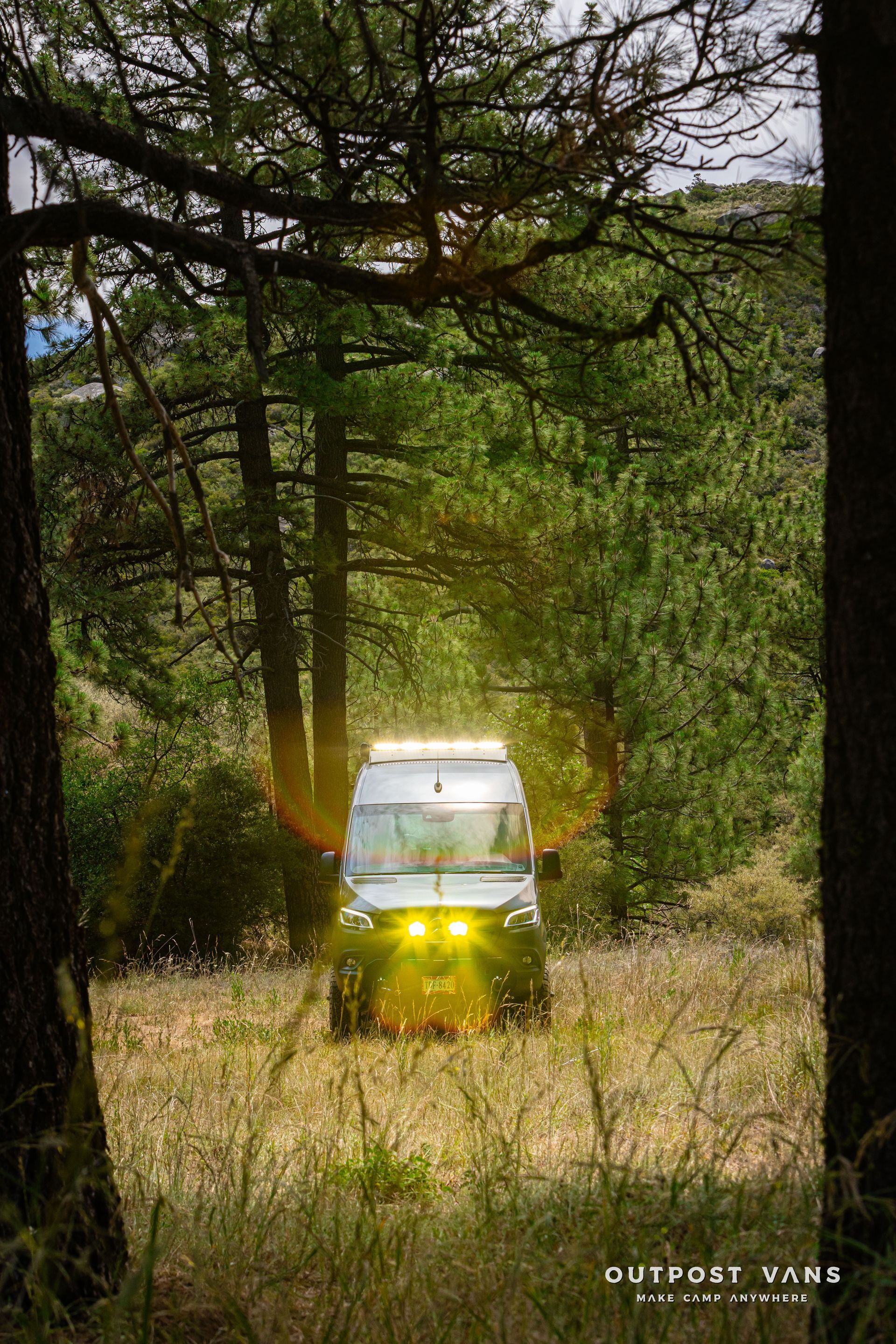 A van is driving through a forest with the sun shining through the trees. Outpost vans Sprinter 170 AWD.