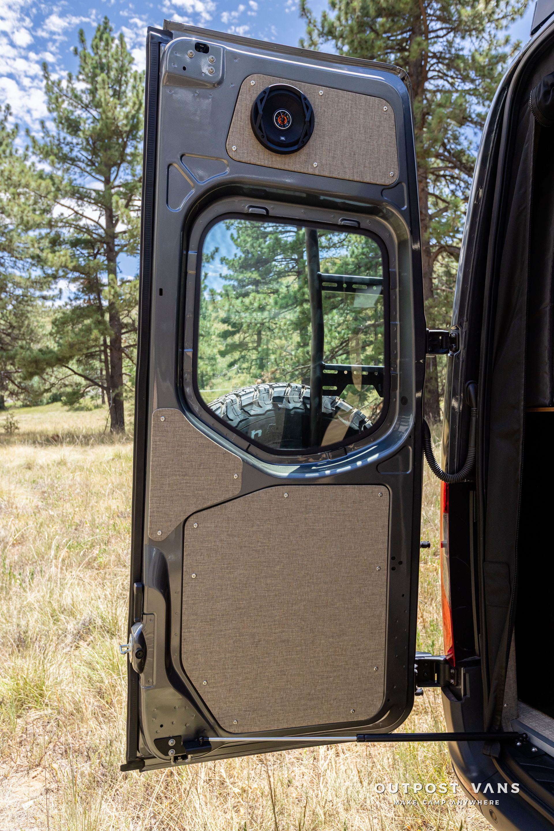 A van with the door open is parked in a field with trees in the background. Outpost vans Sprinter 170 AWD.