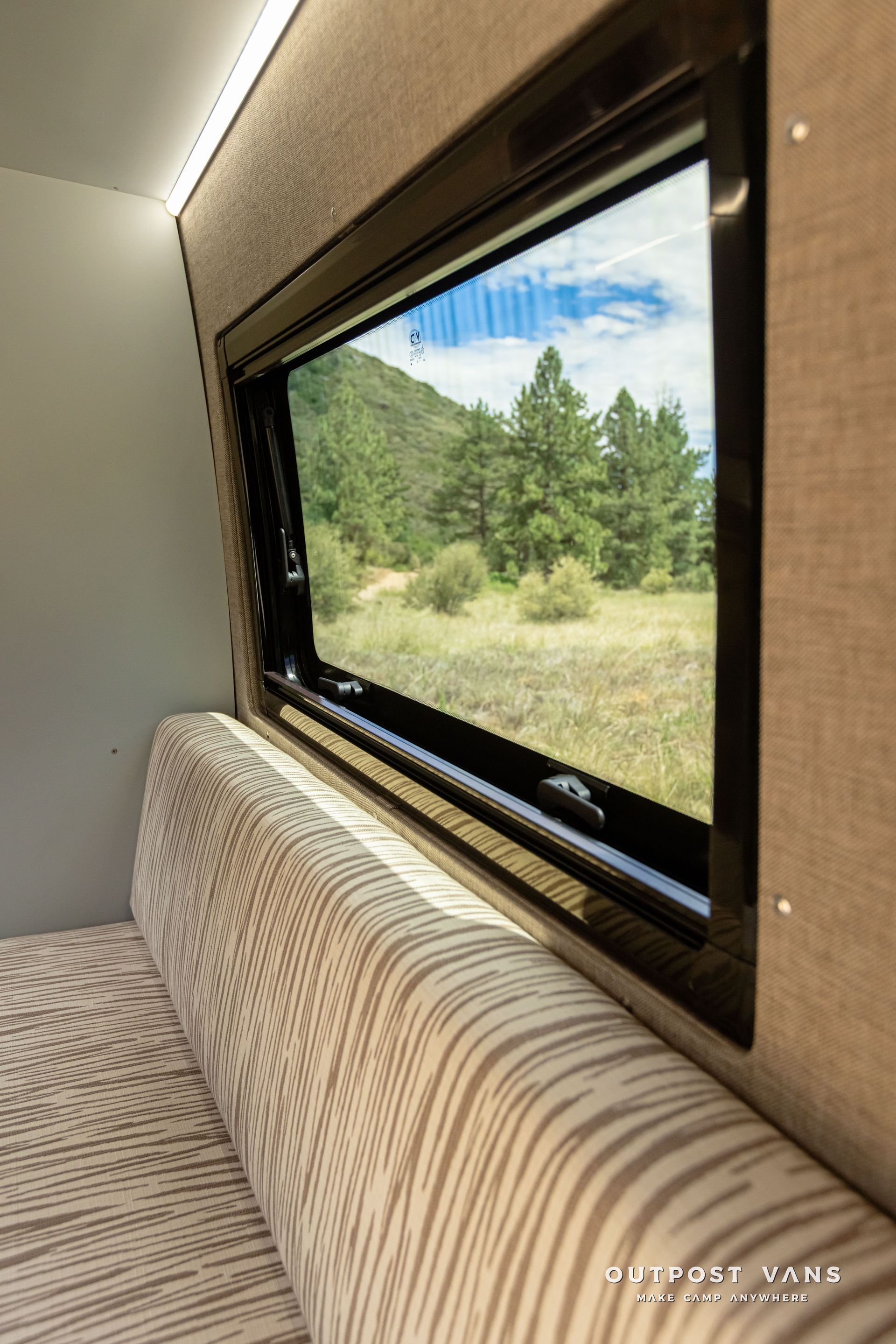 A couch is sitting next to a window with a view of a field. Outpost vans Sprinter 170 AWD.