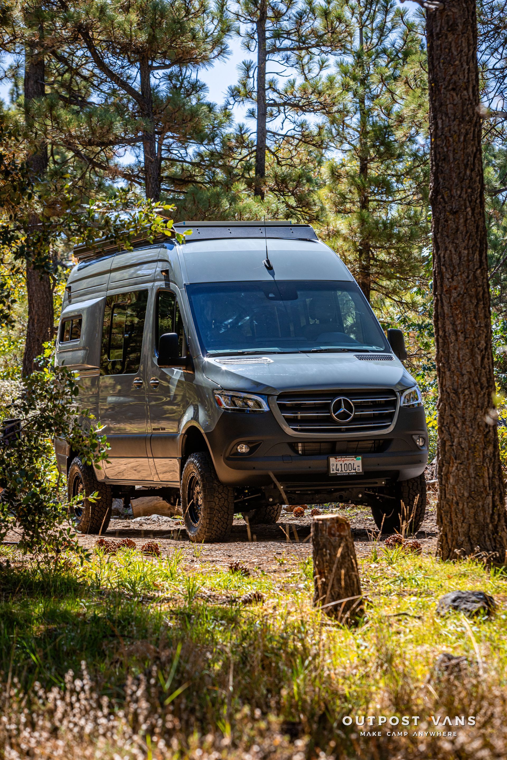 Sprinter camper van parked on a dirt road in a sunny forest, surrounded by trees.
