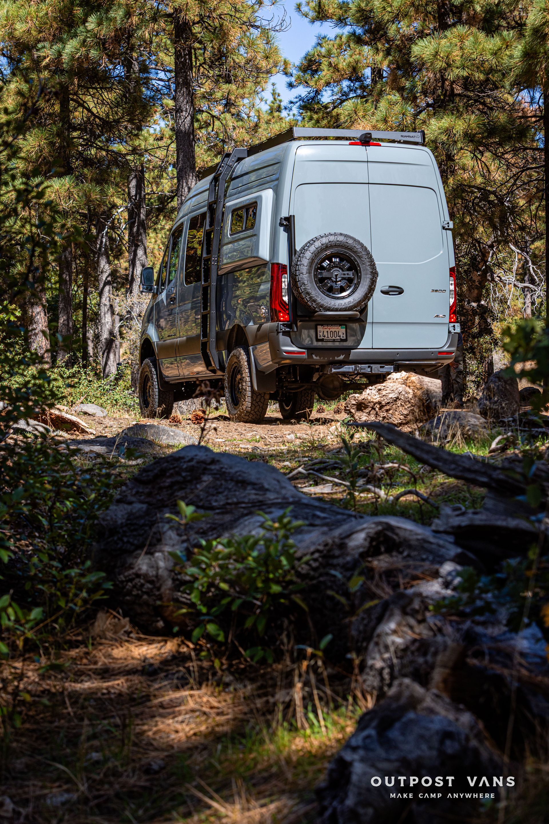 A Sprinter light-colored camper van drives down a dirt road through a forest.