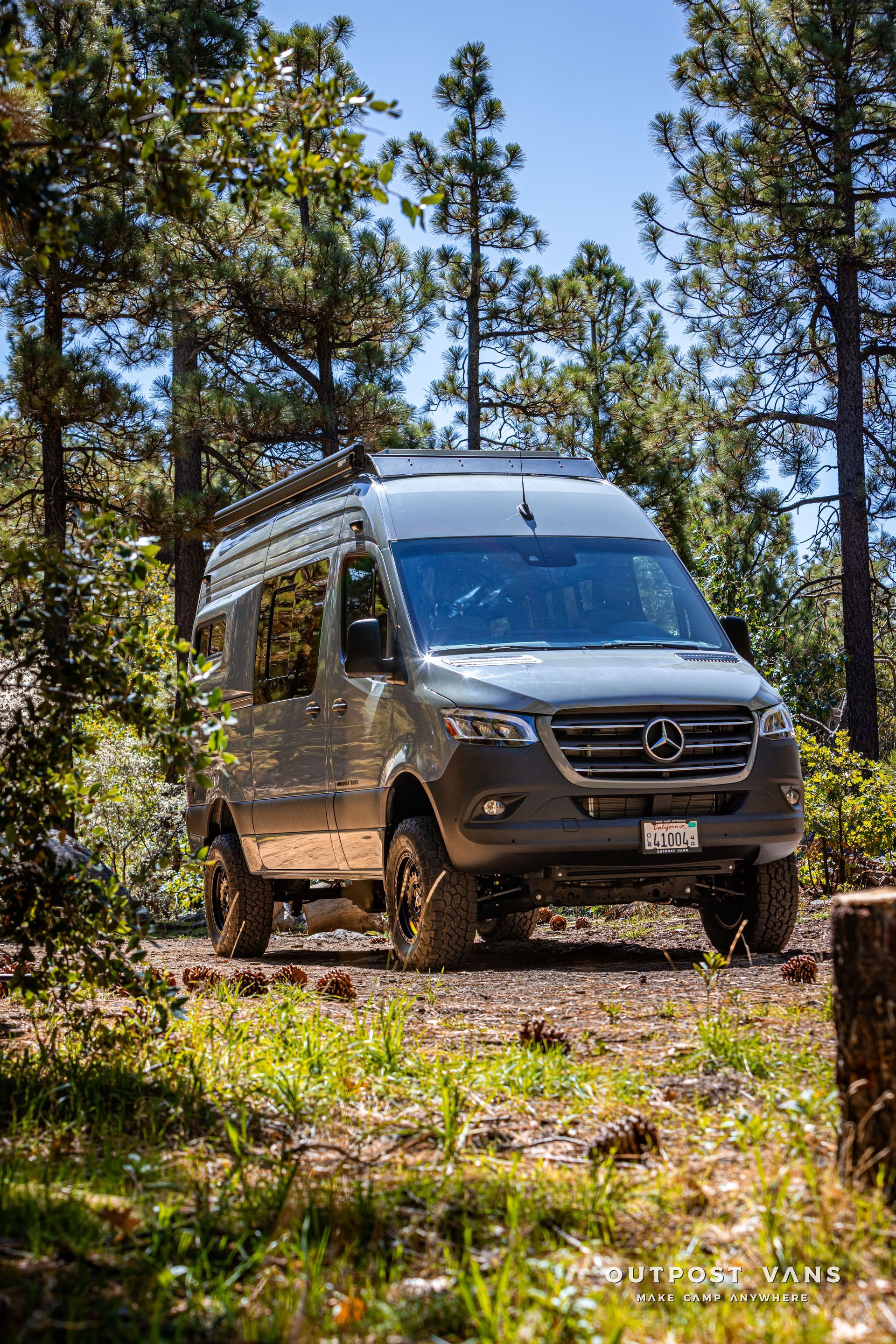 Sprinter Grey Mercedes Sprinter van with off-road tires parked on a dirt path in a forest.