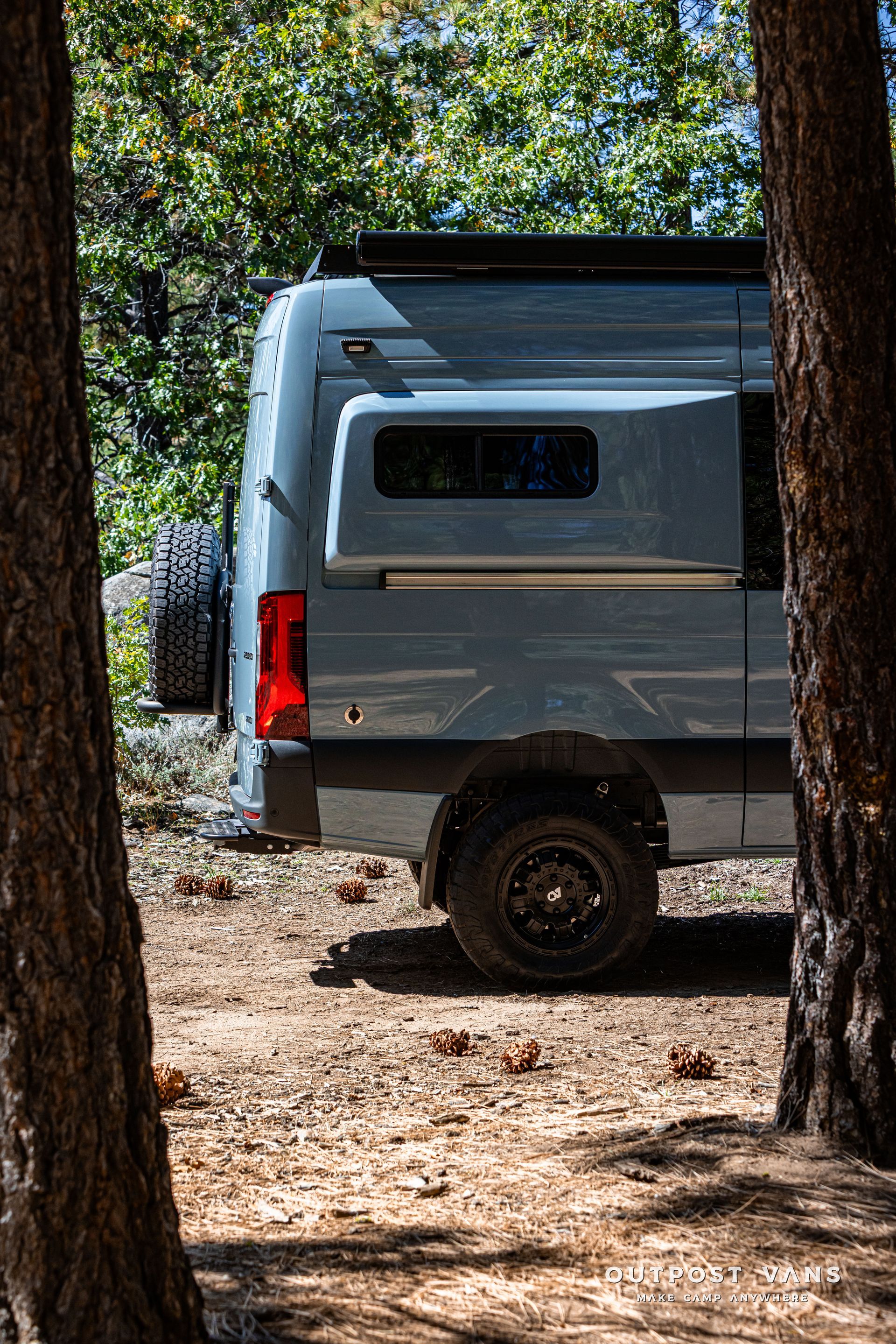 Sprinter Blue camper van parked in a forest, viewed between tree trunks.