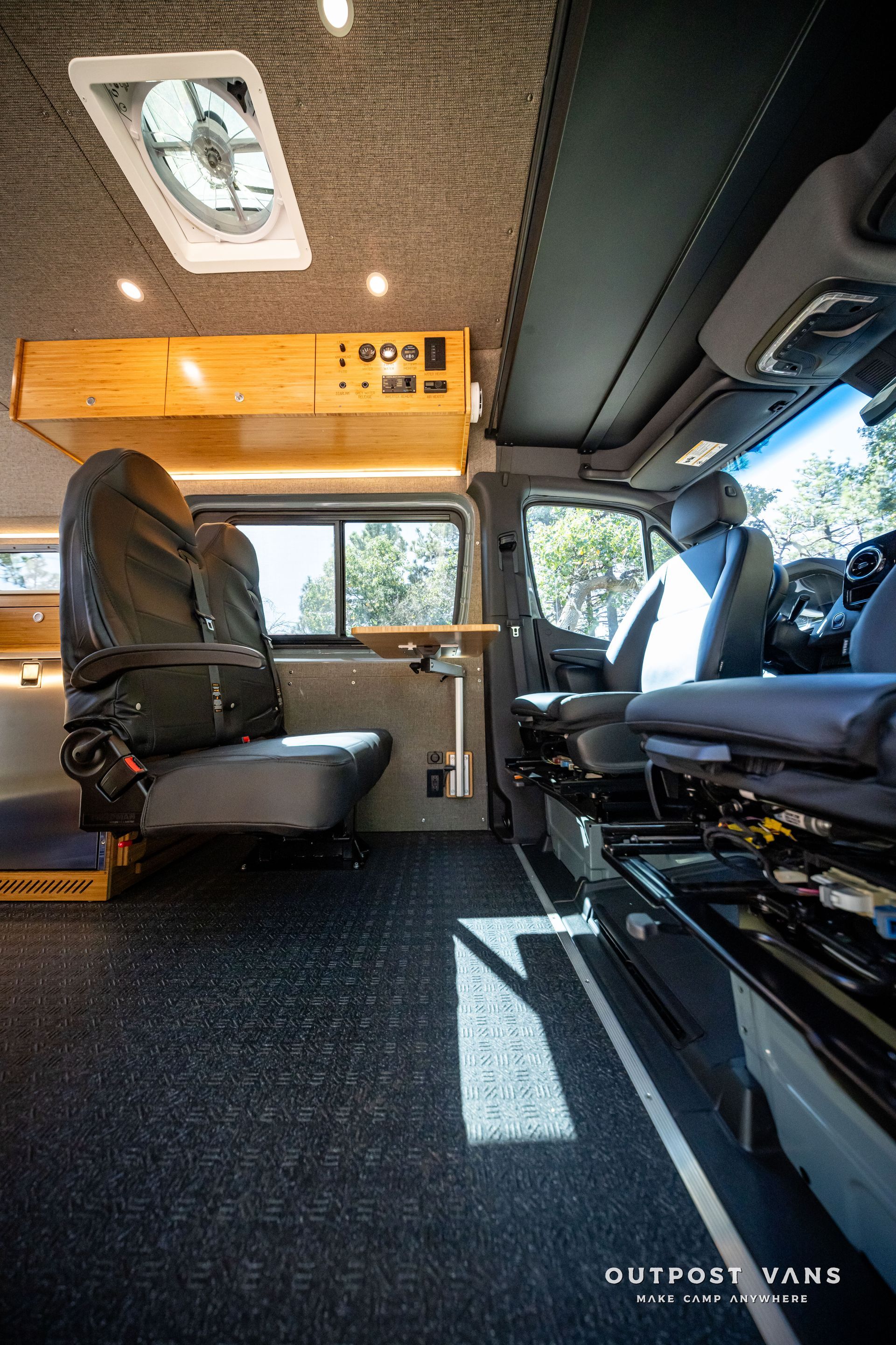 Sprinter Interior of a camper van with dark gray floor and seats. A window shows sunlight. Ceiling fan and wood accents.