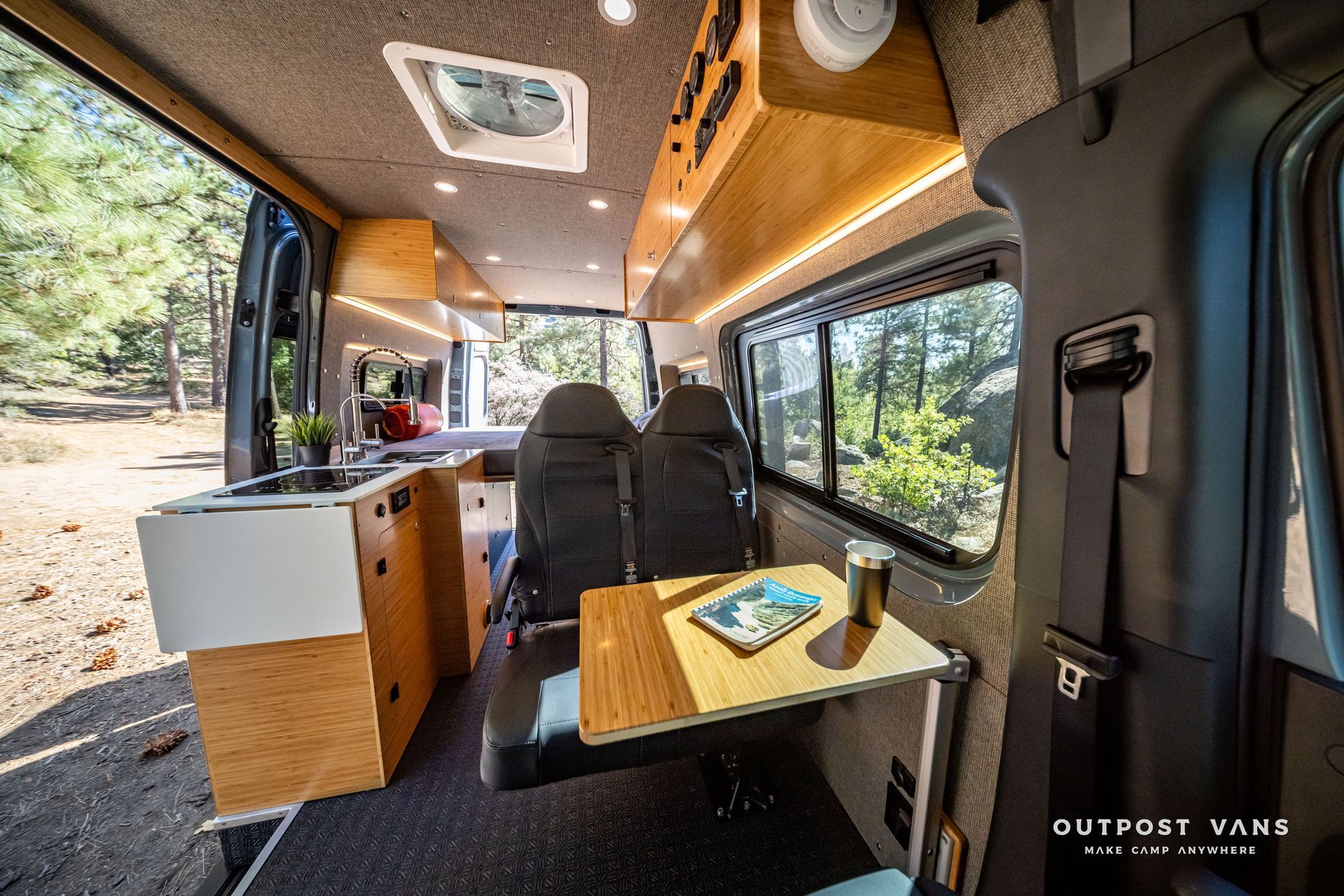 Sprinter Interior of a van with a wooden kitchenette, table, and seats, parked in a wooded area.
