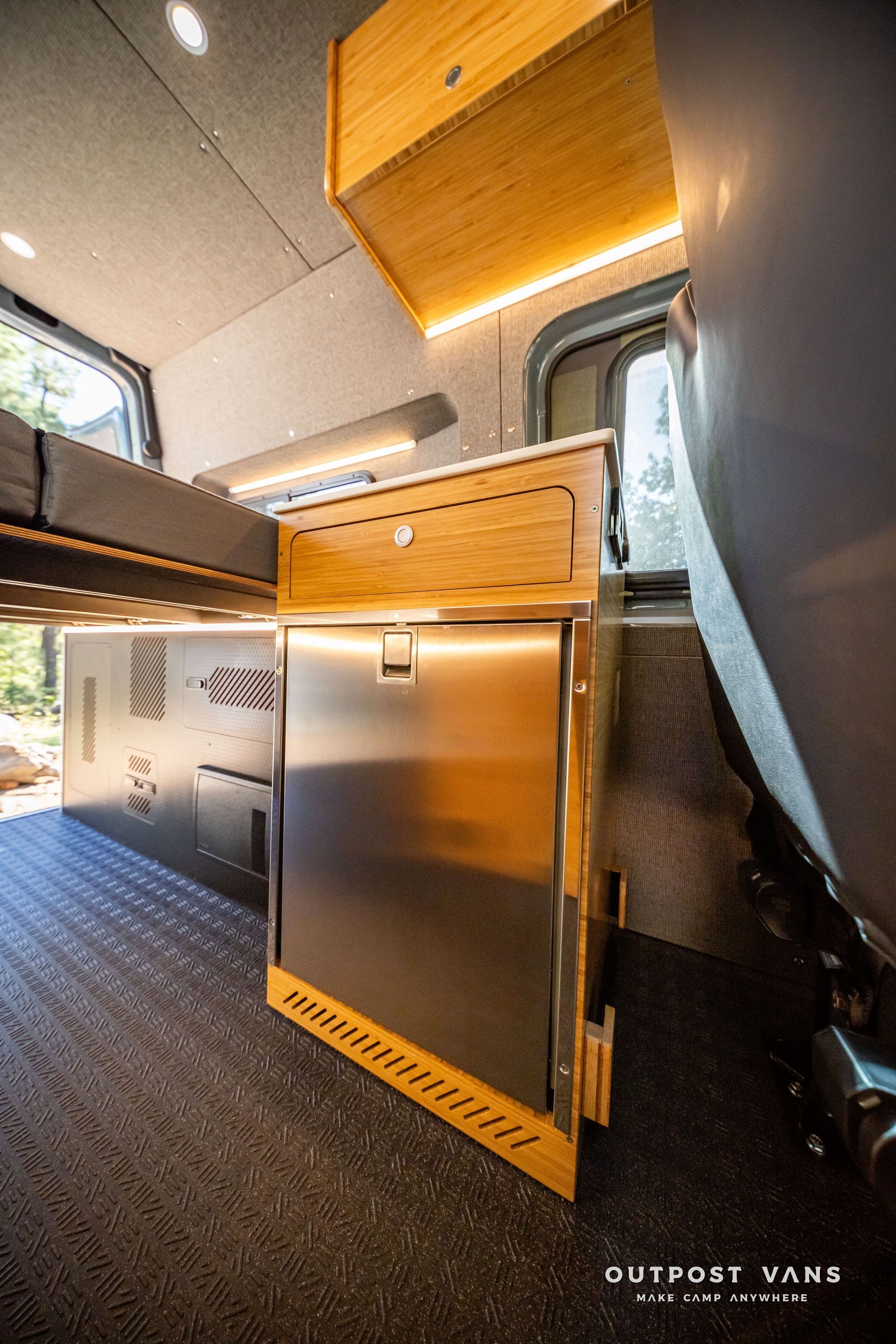 Sprinter Inside view of a camper van with a stainless steel refrigerator, wooden cabinets, and overhead lighting.